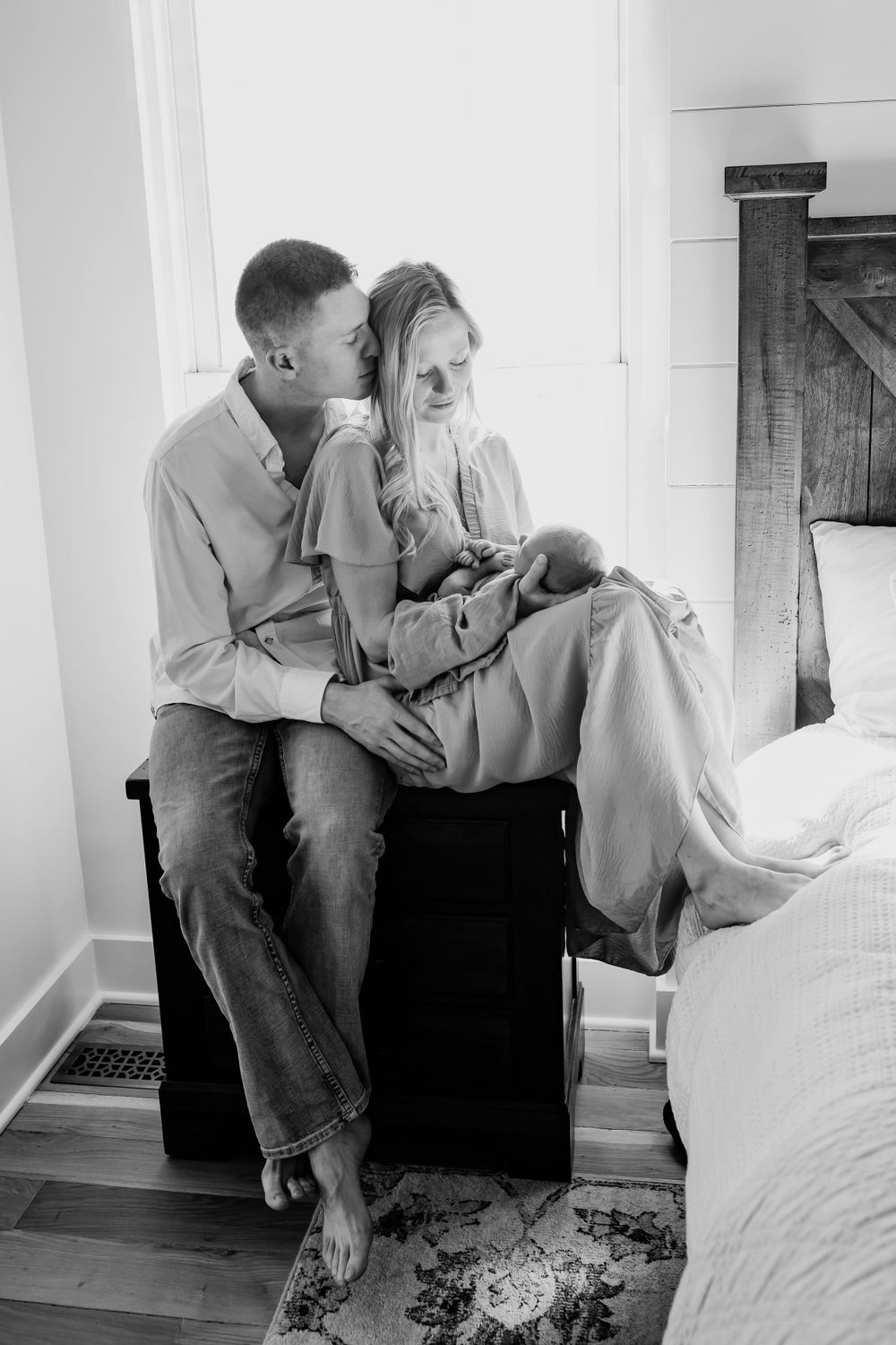 A tender moment captured in black and white as a couple sits together intimately on a wooden chest in a bedroom setting.