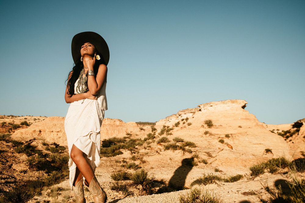 A couple in white outfits poses intimately against a scenic desert landscape during golden hour.