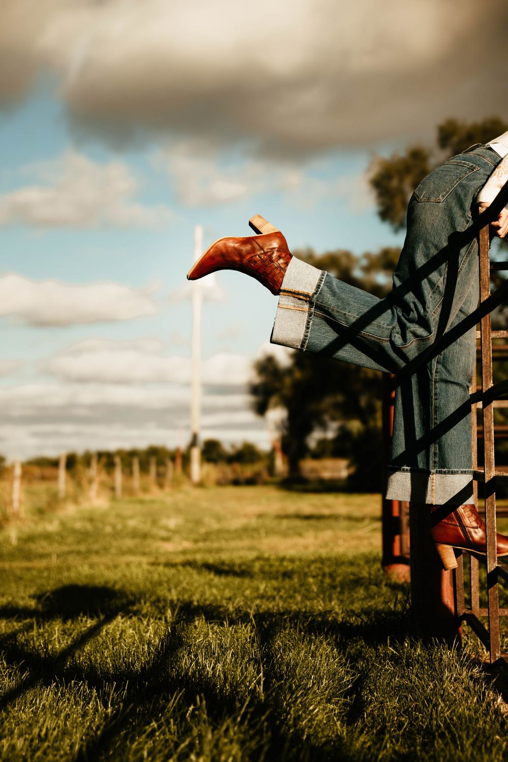 A leather boot hangs from a metal gate in a rural field at sunset with dramatic clouds in the background.