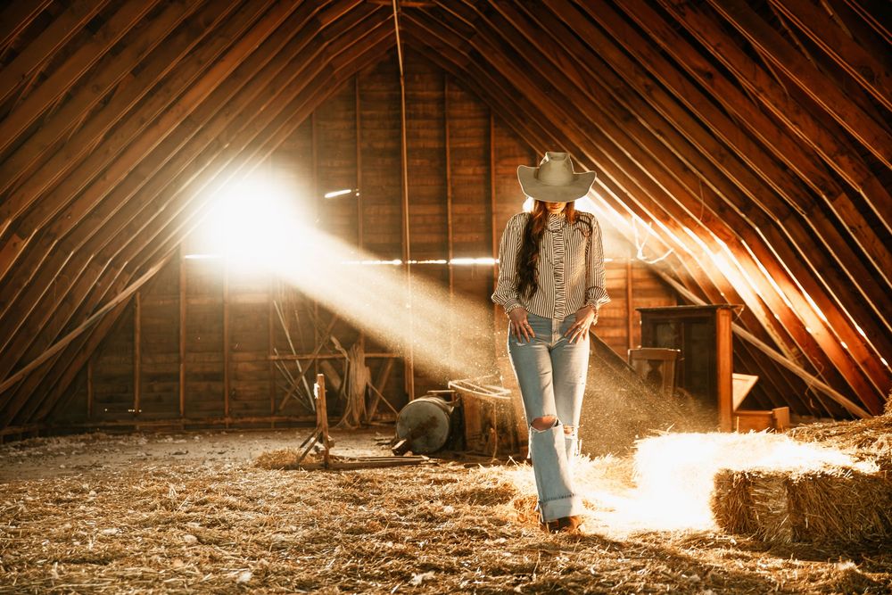 A figure stands in a sunlit attic or barn loft with dramatic rays of light streaming through the wooden beams.