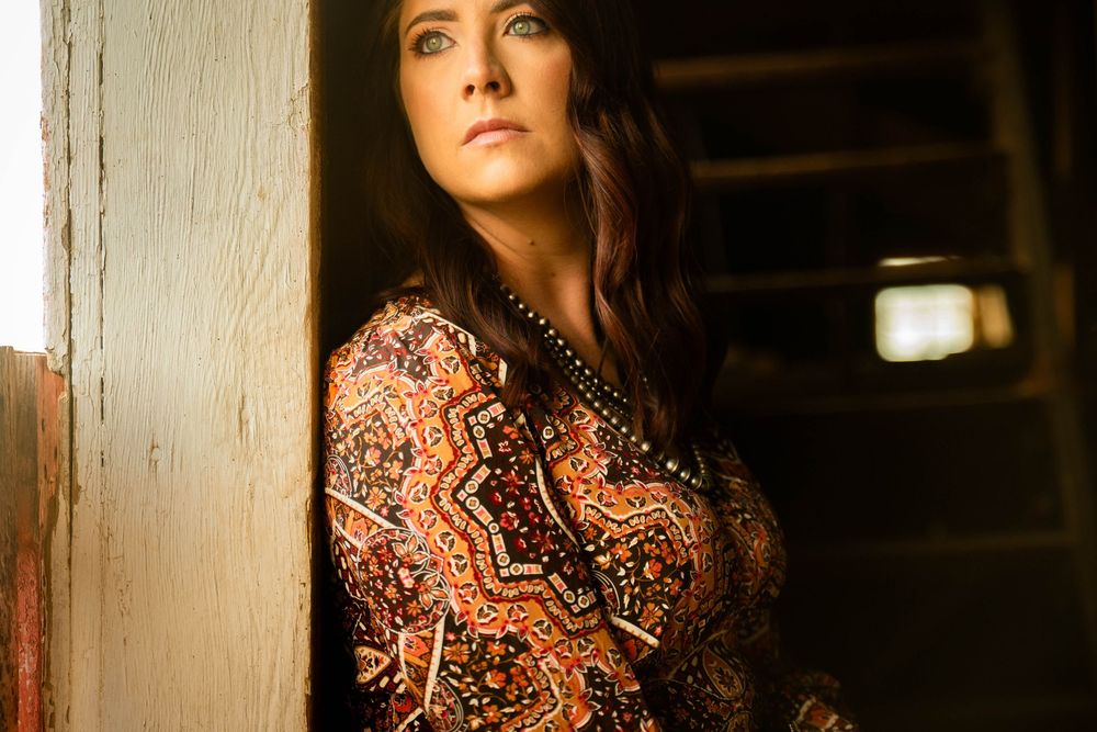 A person in a floral print dress poses against a rustic wooden wall in dramatic lighting.