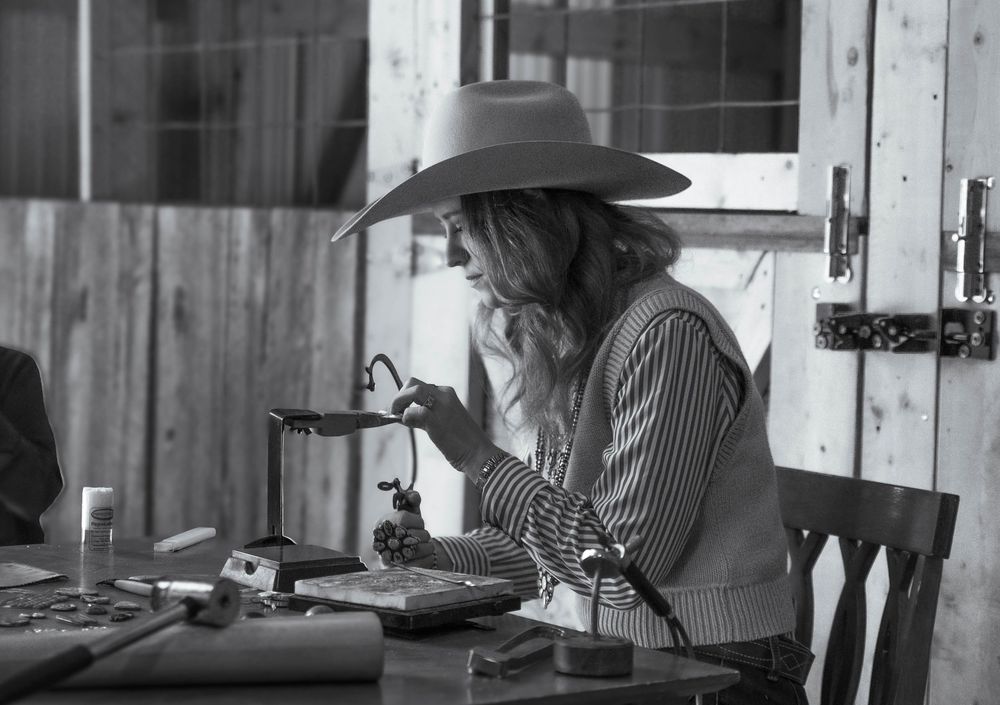A black and white photograph shows a person in a hat working at a table in a rustic setting.