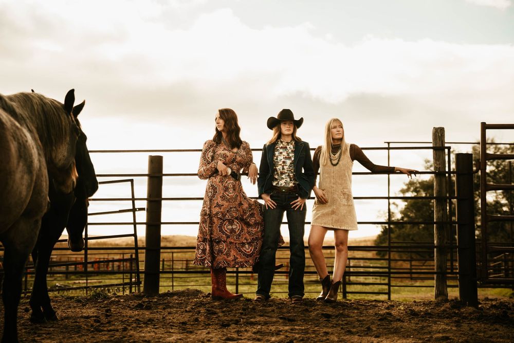 Three people in western attire stand by a metal gate at sunset with a horse silhouetted in the foreground.