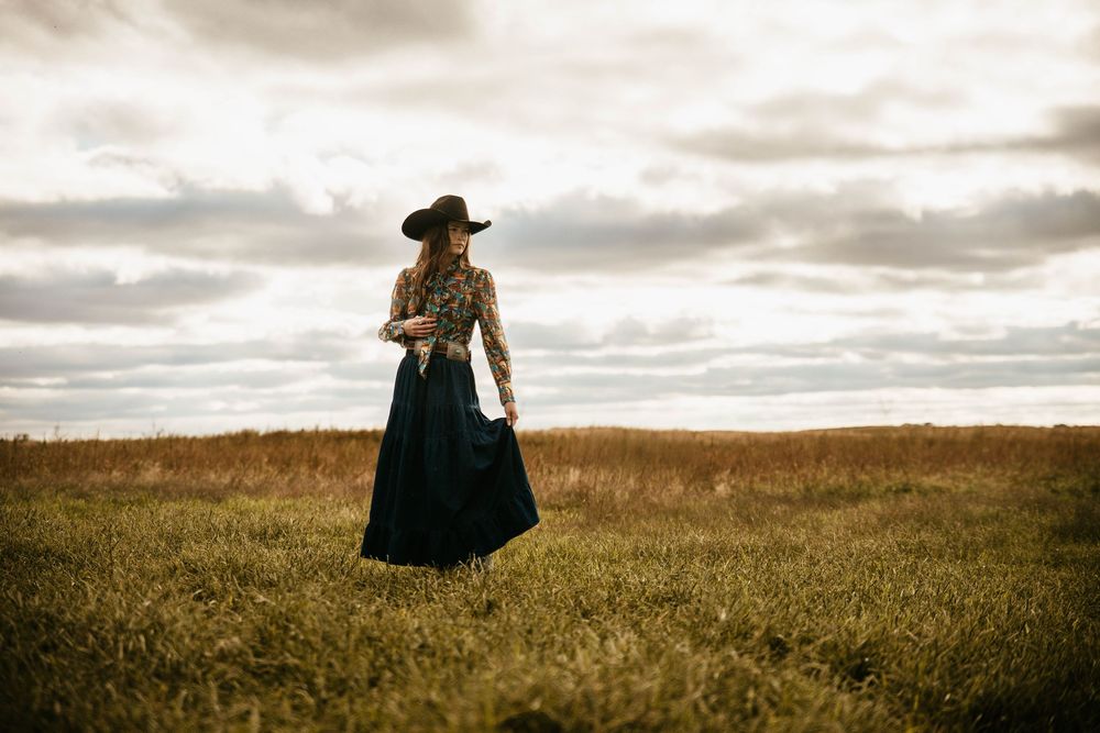 A person in dark clothing stands in a golden field beneath moody clouds at sunset.