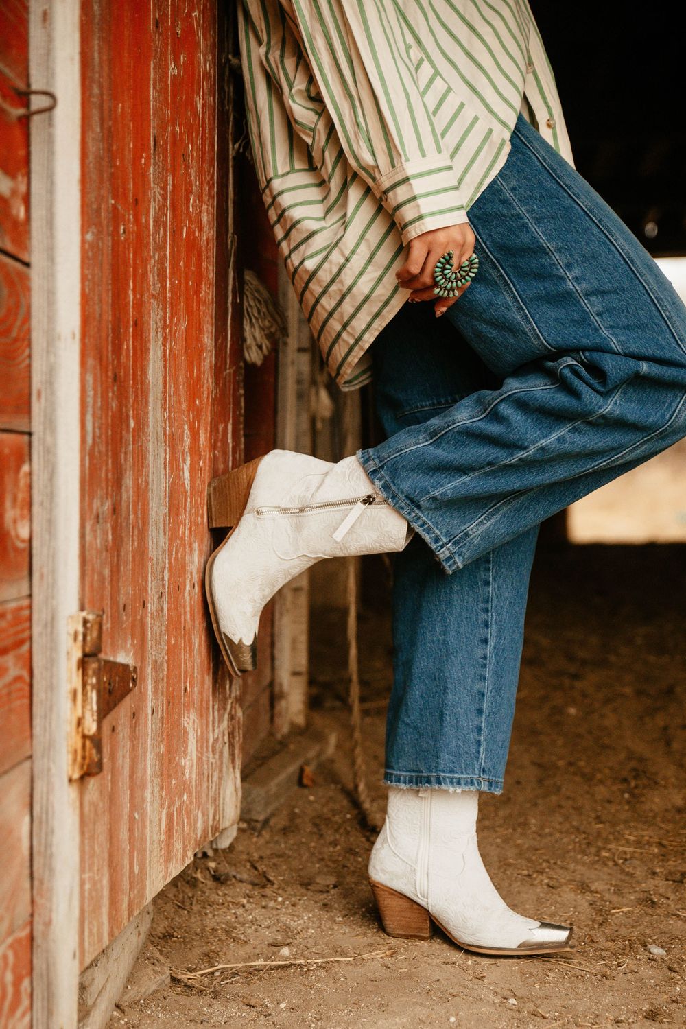 Person wearing white cowboy boots and blue jeans steps through a rustic red doorway.