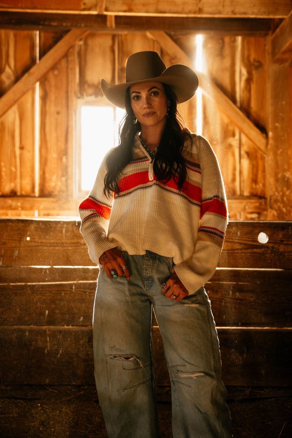 Western style portrait in a rustic barn featuring a striped sweater, cowboy hat and distressed jeans in warm lighting.