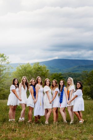 Nine women in white dresses with graduation sashes pose together in a scenic mountain meadow under cloudy skies.