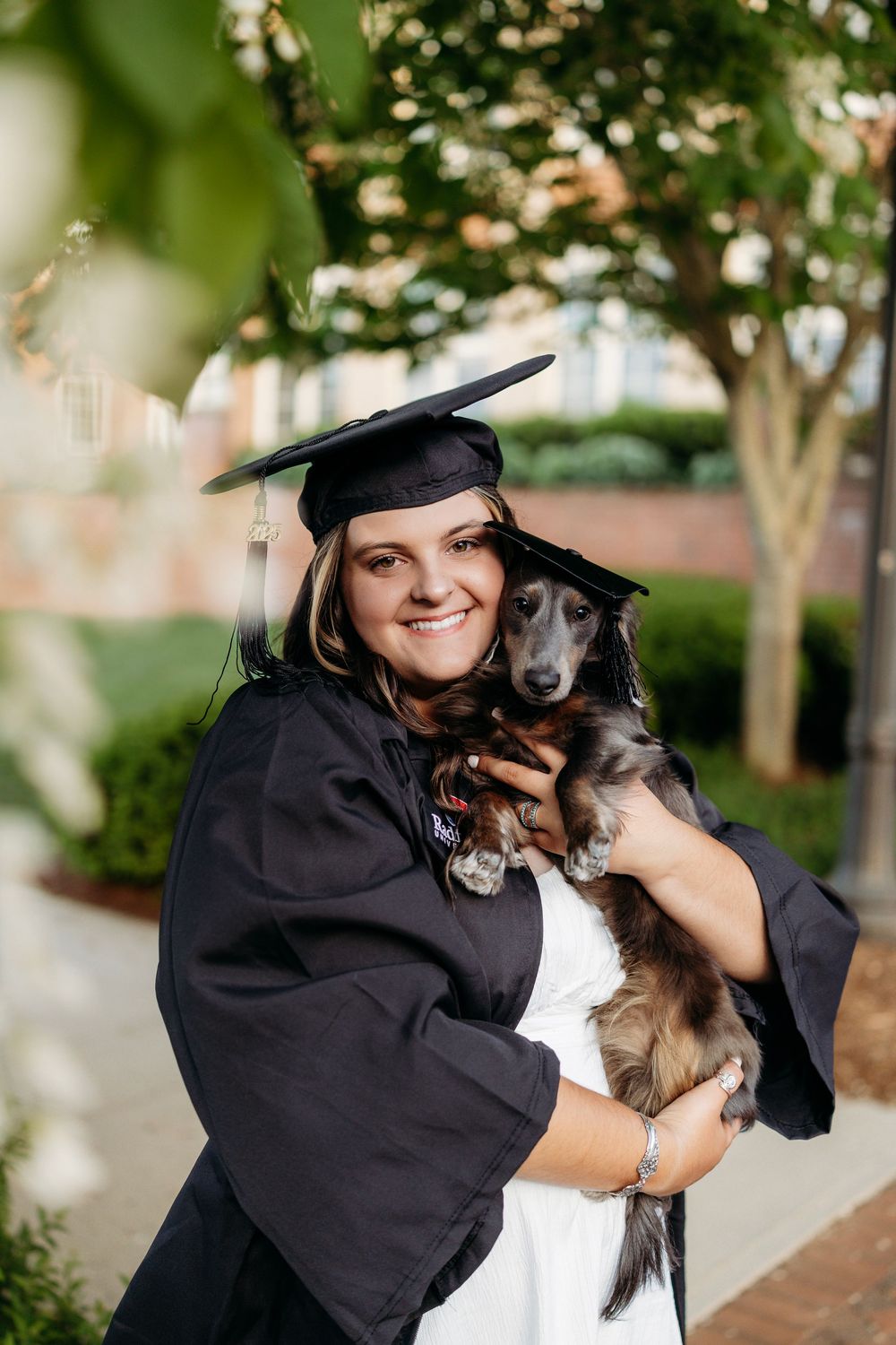 Smiling graduate in black cap and gown holds a long-haired dachshund outdoors on a college campus.