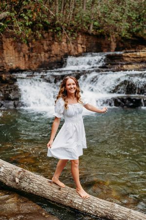 Young woman in white off-shoulder dress balances on a log over a serene river with a waterfall behind her.