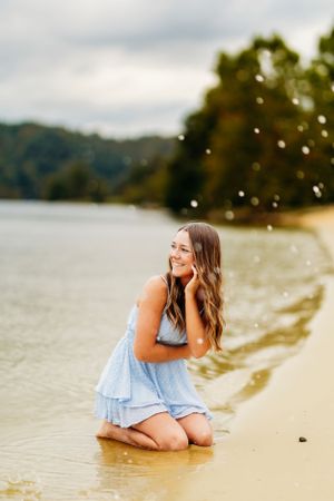 Smiling young woman in blue dress kneeling at lake shore with bubbles in cloudy sky.