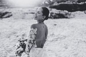 A joyful bride in a backless wedding dress holds a bouquet while laughing in black and white photography.