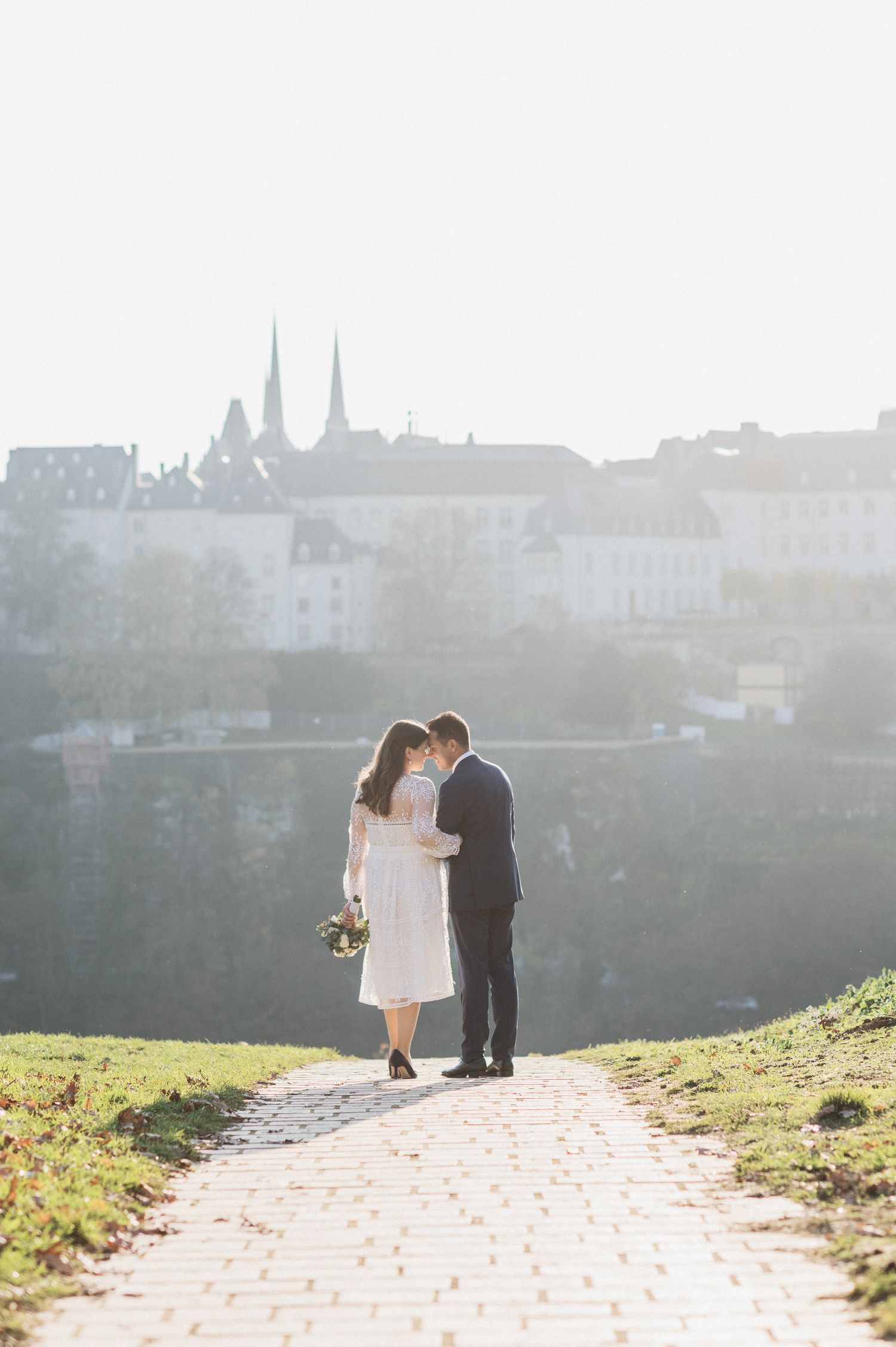 A couple shares an intimate moment on a cobblestone path overlooking a misty European cityscape with church spires.