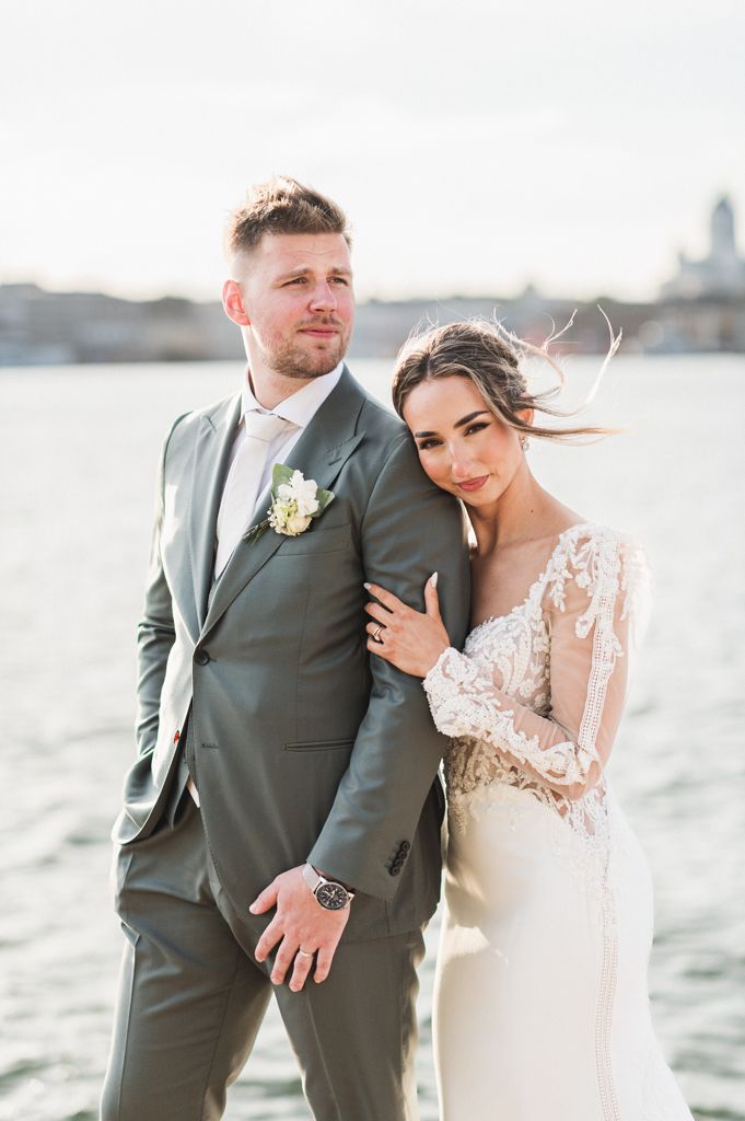 Newlywed couple poses together by waterfront, bride in lace gown and groom in gray suit on sunny day.