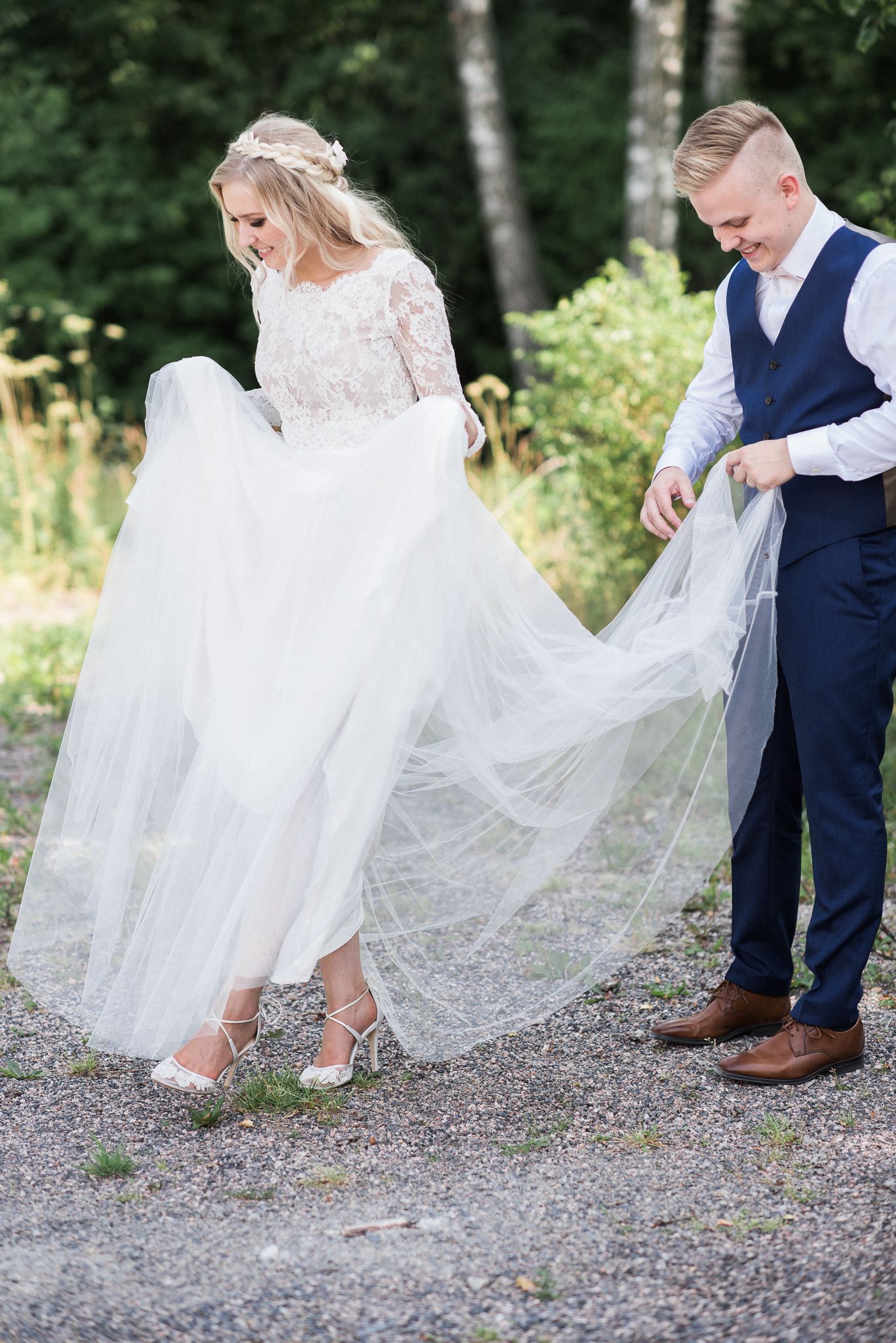A bride in a lace dress lifts her gown while walking on a forest path as her partner helps with the flowing veil.