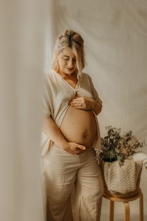 Expectant mother in soft beige clothing stands by a rustic wooden stool with dried flowers in a warm, dreamy setting.