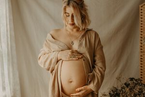 A maternity photo in warm neutral tones shows a pregnant woman wearing an oversized beige sweater against a light backdrop.