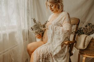 Ethereal vintage-style portrait in warm tones showing a pregnant in white lace dress seated by window with dried flowers.