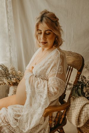 Woman in white lace robe sits in wooden chair surrounded by dried flowers in warm vintage-style boudoir photo. Maternity photography
