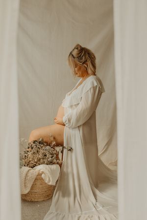 Expectant mother in flowing white robe stands beside woven basket in ethereal maternity photo against pale backdrop. Beckenham photography