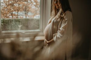 Silhouette of pregnant woman in white robe standing by window during autumn day.