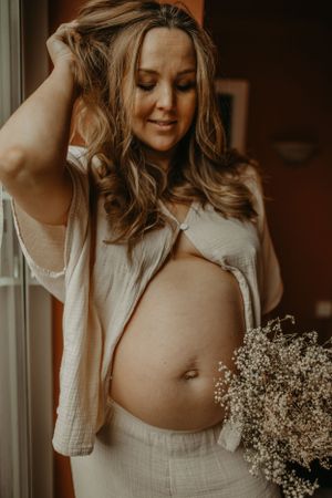 A pregnant woman in a white outfit poses with baby's breath flowers in a warm, moody setting.