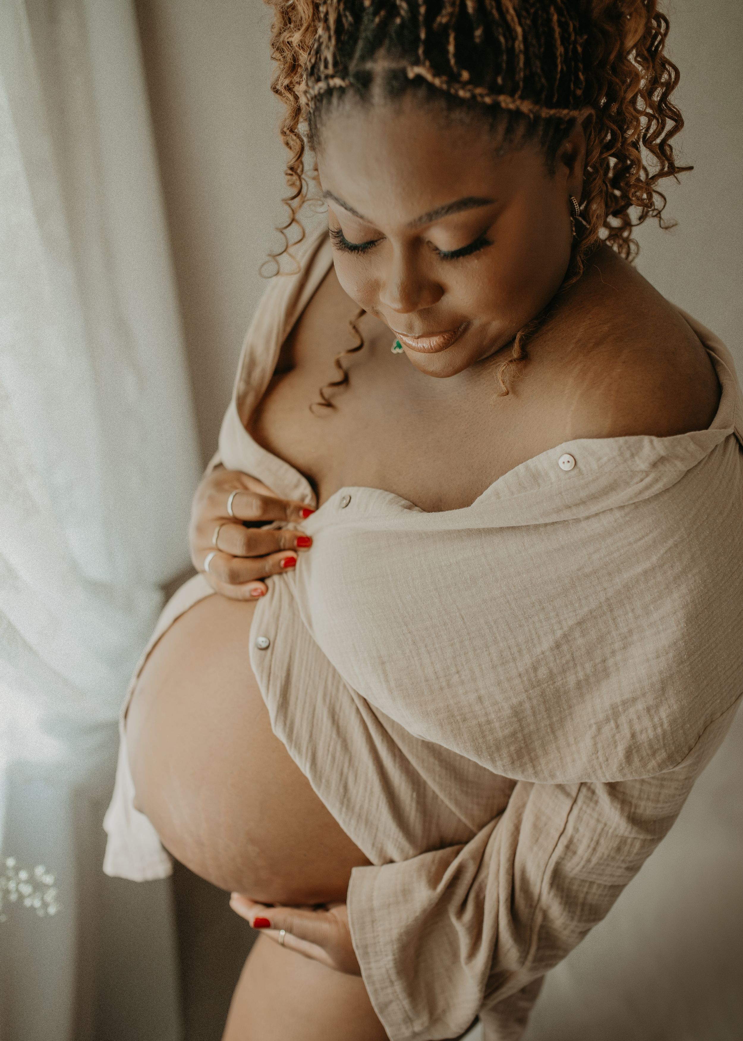 A pregnant woman in a cream colored off-shoulder dress poses for a maternity photo in soft natural lighting.