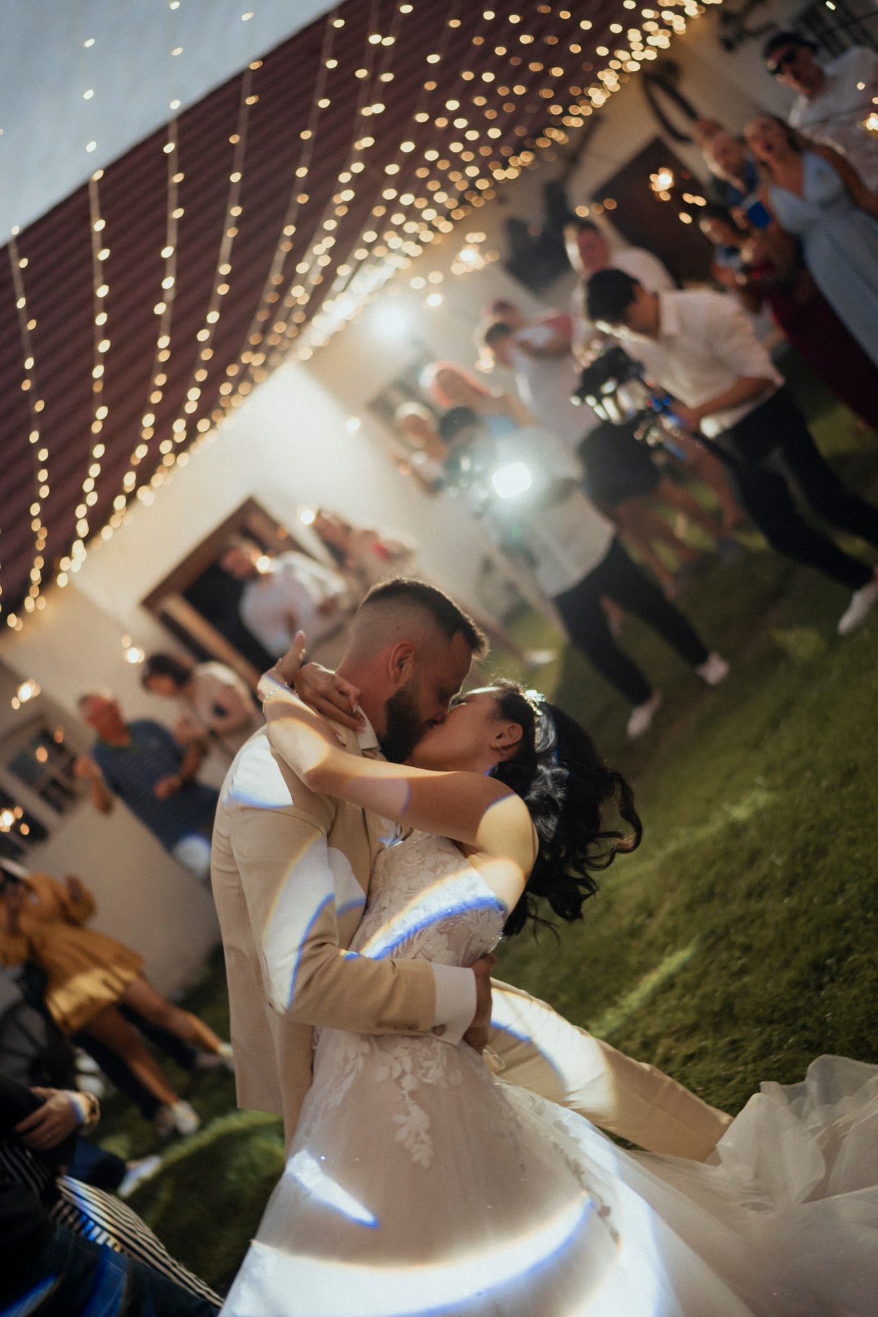 Romantic wedding dance under glowing string lights with guests gathering around.
