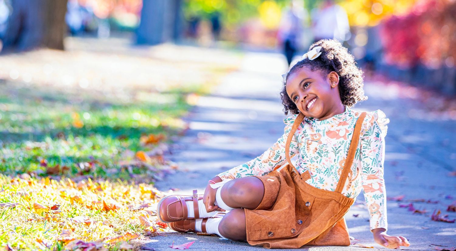 Young child sits on autumn leaves wearing floral dress and brown overalls with joyful smile.