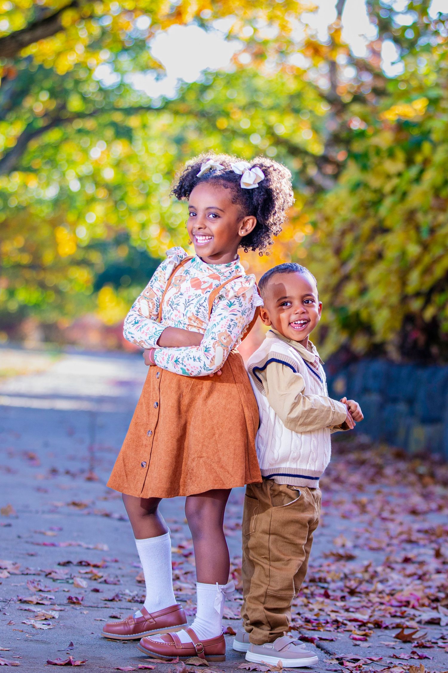 Two children pose playfully among fall foliage on a leaf-covered path.