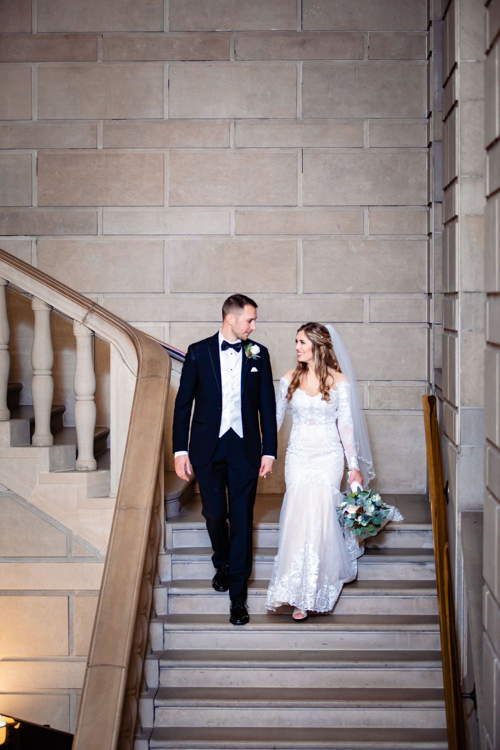 Bride and groom walking down elegant stone staircase, she in lace gown with veil, he in black tuxedo with bow tie.