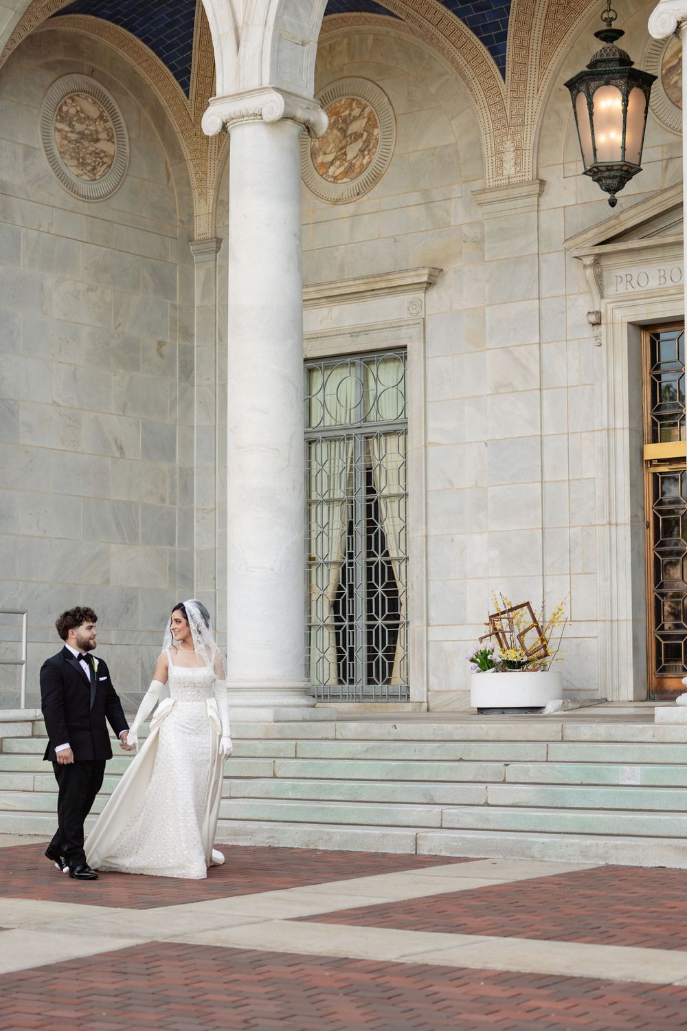 Bride and groom walk hand in hand outside an elegant neoclassical building with marble columns and arched entryway.