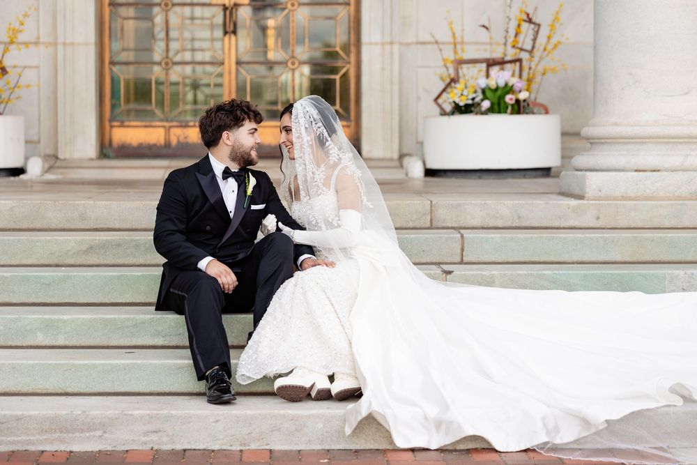 Bride and groom sit on stone steps sharing a romantic moment, bride in flowing white gown with veil, groom in black tuxedo.