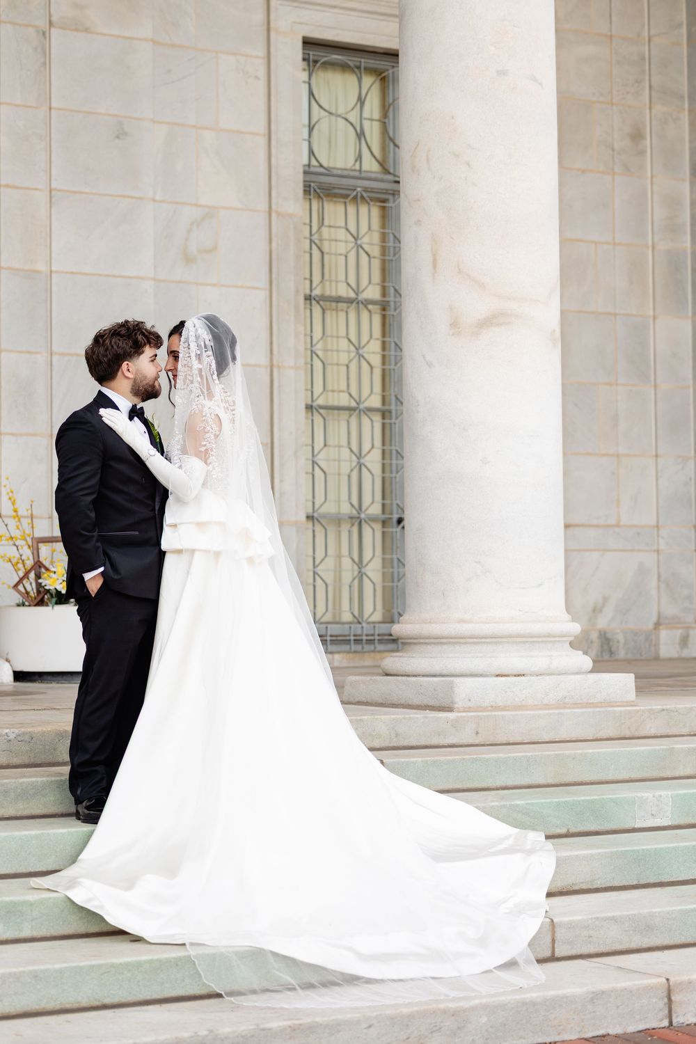 Bride in white gown with veil and groom in black tuxedo share a romantic moment on marble steps.