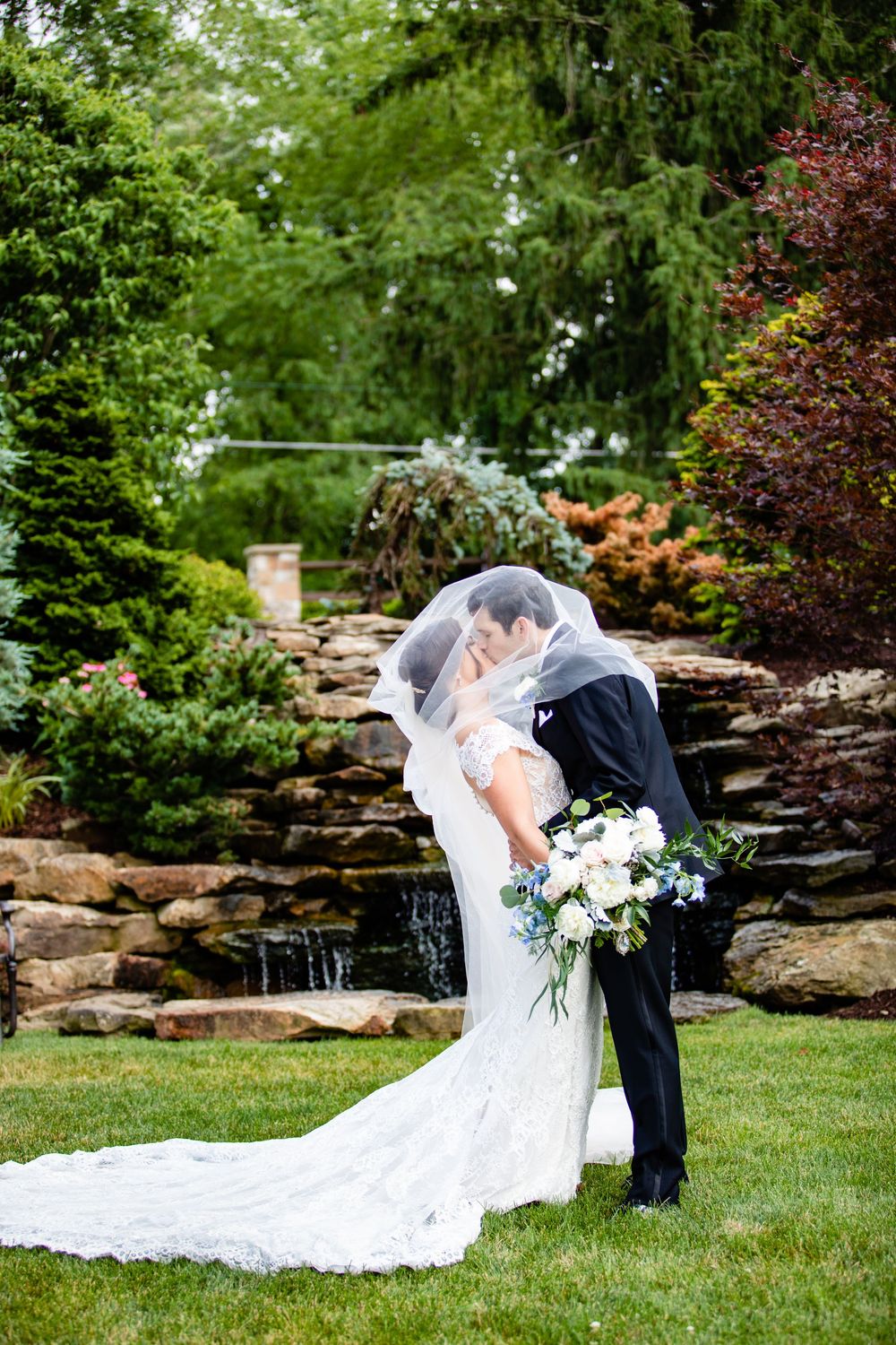 Bride and groom share a romantic kiss under a veil in a lush garden with a stone waterfall backdrop.