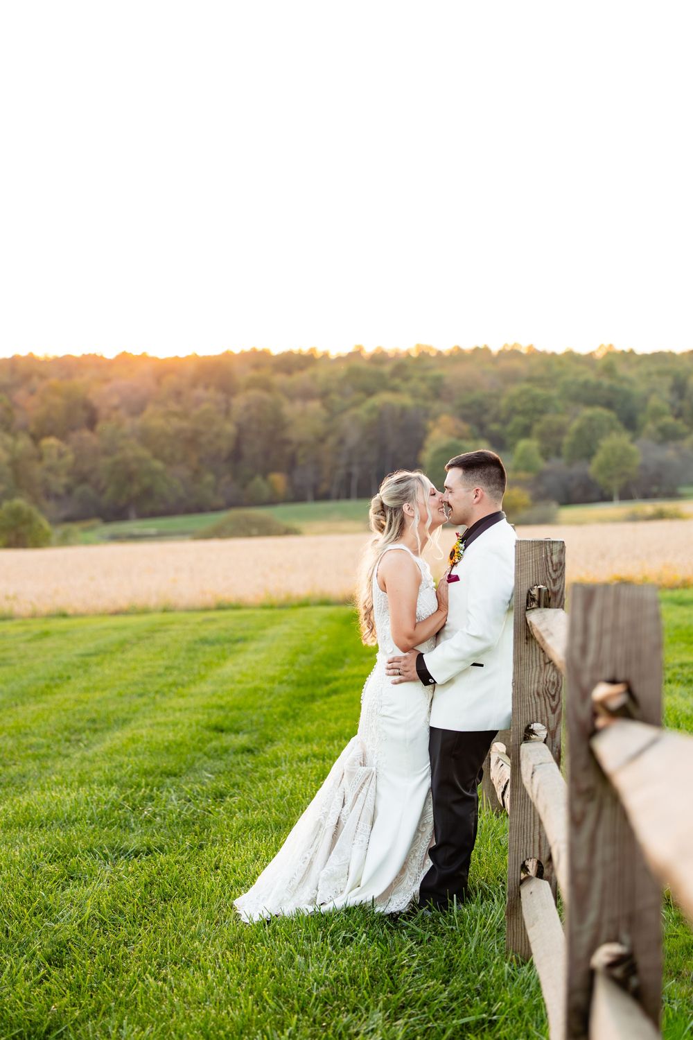 Bride and groom share a romantic moment by a rustic fence at golden hour, surrounded by lush green fields.