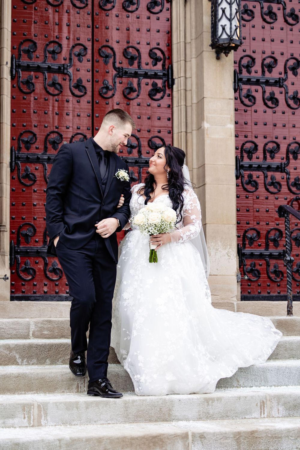 Bride and groom share a romantic moment on church steps, she in lace gown with bouquet, he in black suit.