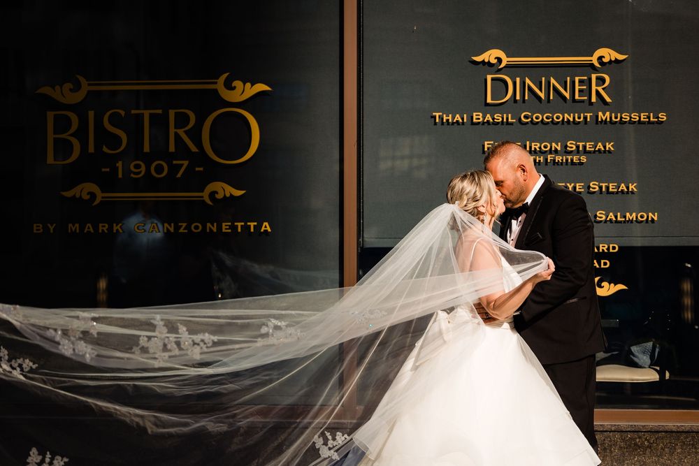 Bride and groom share romantic kiss outside Bistro 1907 restaurant, wedding veil flowing dramatically in the wind.
