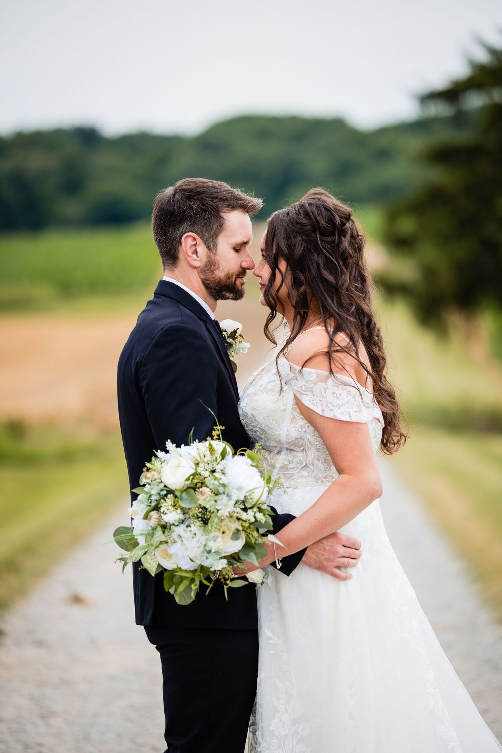 Bride and groom sharing a romantic moment on a country path, with a white floral bouquet and lush green fields behind them.