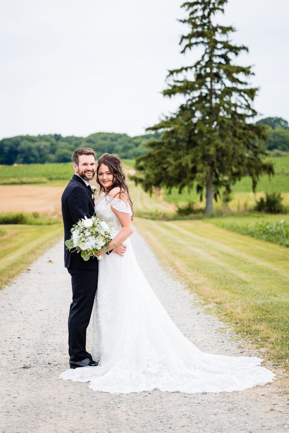Bride and groom pose on a gravel path surrounded by green fields and a tall evergreen tree on their wedding day.