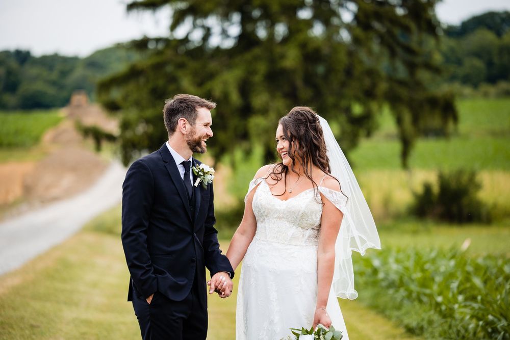 A bride and groom hold hands and smile at each other in a lush green countryside setting on their wedding day.
