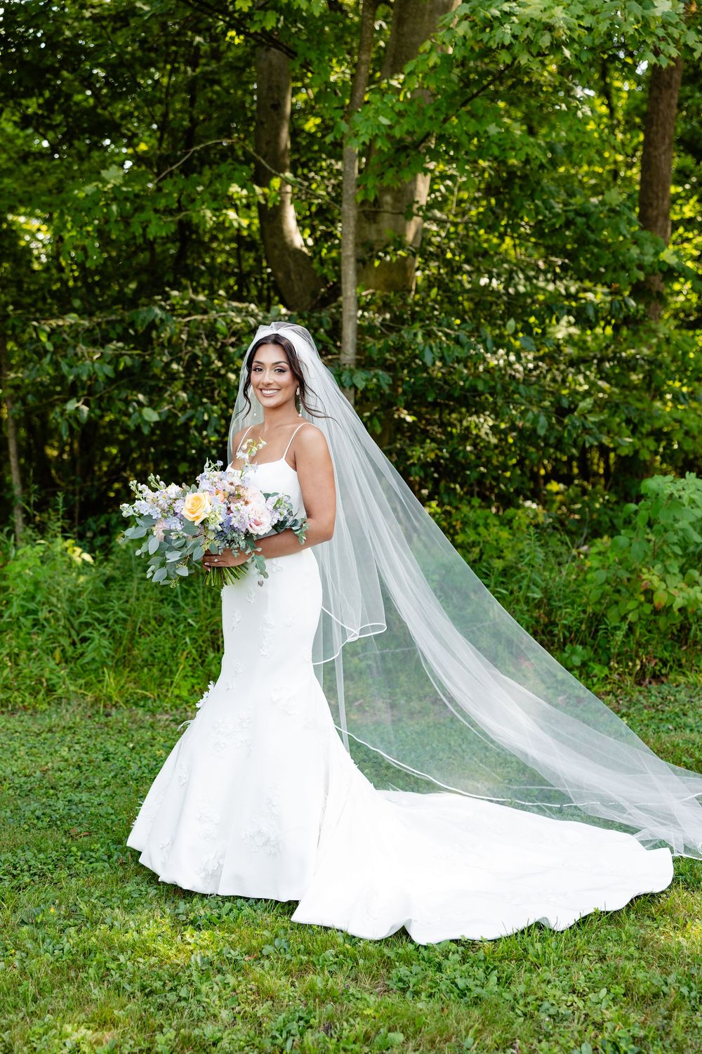 Bride in a white mermaid gown with a long veil holds a floral bouquet, posing outdoors in a lush green garden.