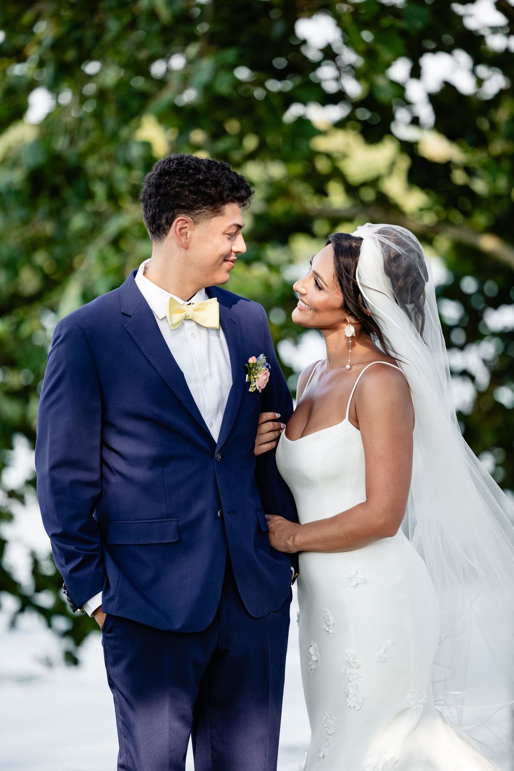 Bride in white gown and veil smiles at groom in navy suit with yellow bow tie outdoors.