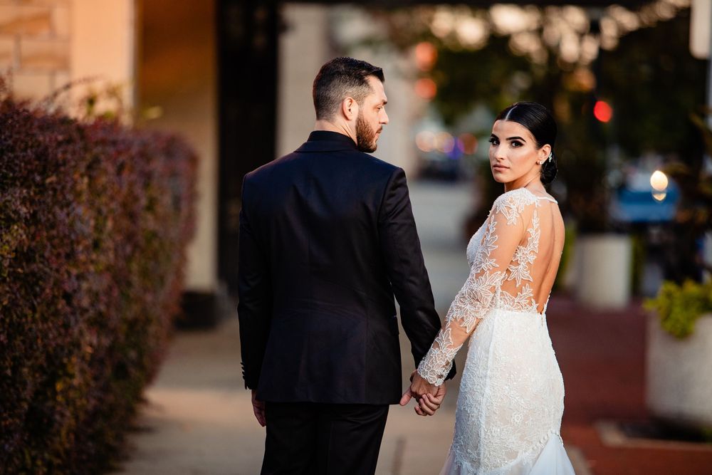 Bride in lace backless gown and groom in black suit hold hands, glancing back at camera during outdoor wedding portrait.