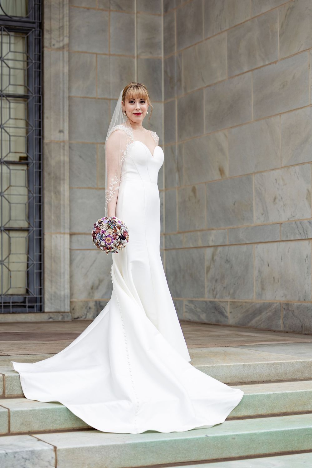 Bride in elegant white mermaid gown with veil holding colorful brooch bouquet on stone steps.