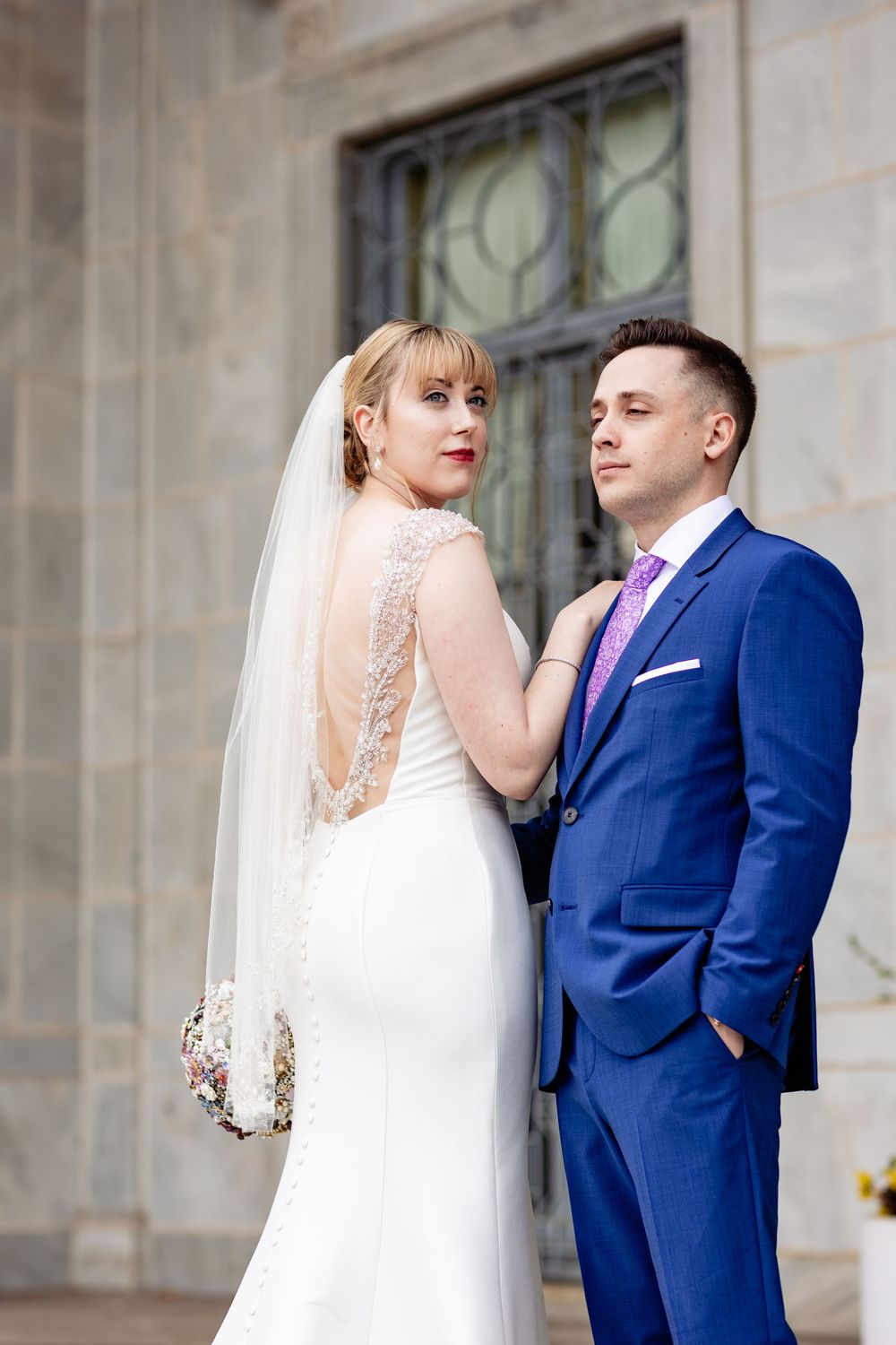 Bride in white lace gown with veil and groom in blue suit pose together outside a stone building.