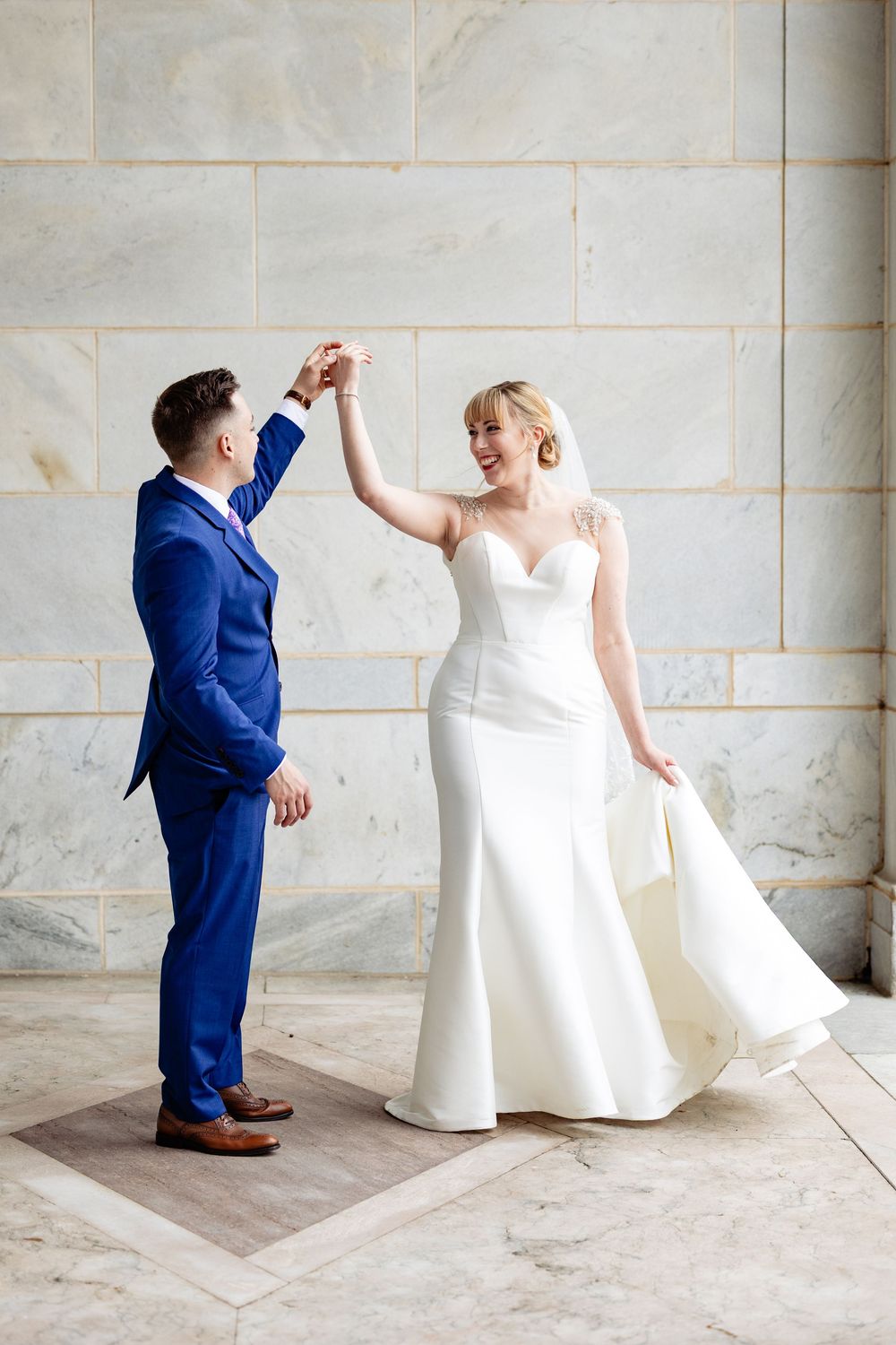 Bride and groom dancing together, she in a white gown, he in a blue suit, against a marble wall.