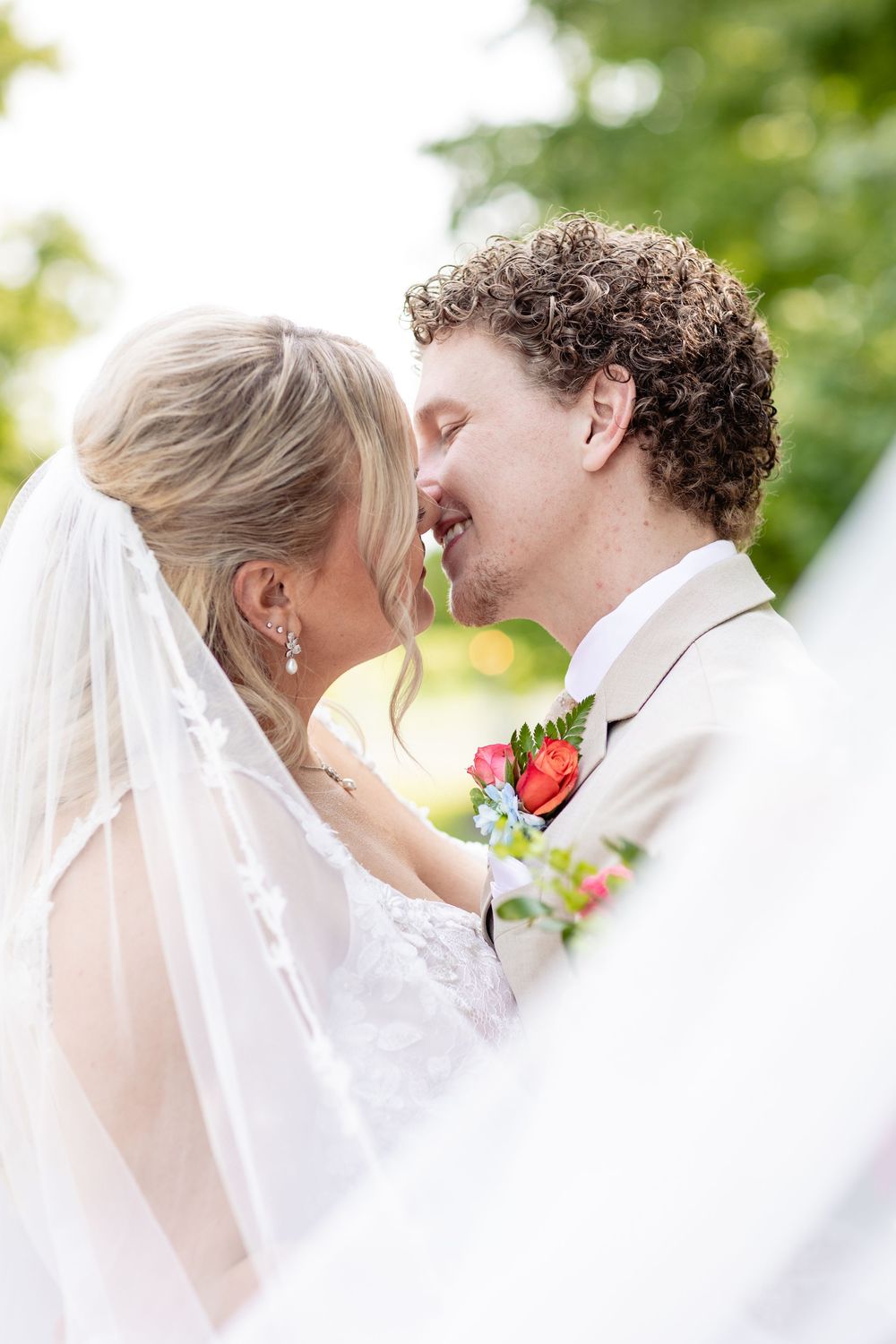 Bride and groom sharing a romantic moment, nearly kissing, with veil draped around them and floral boutonniere outdoors.