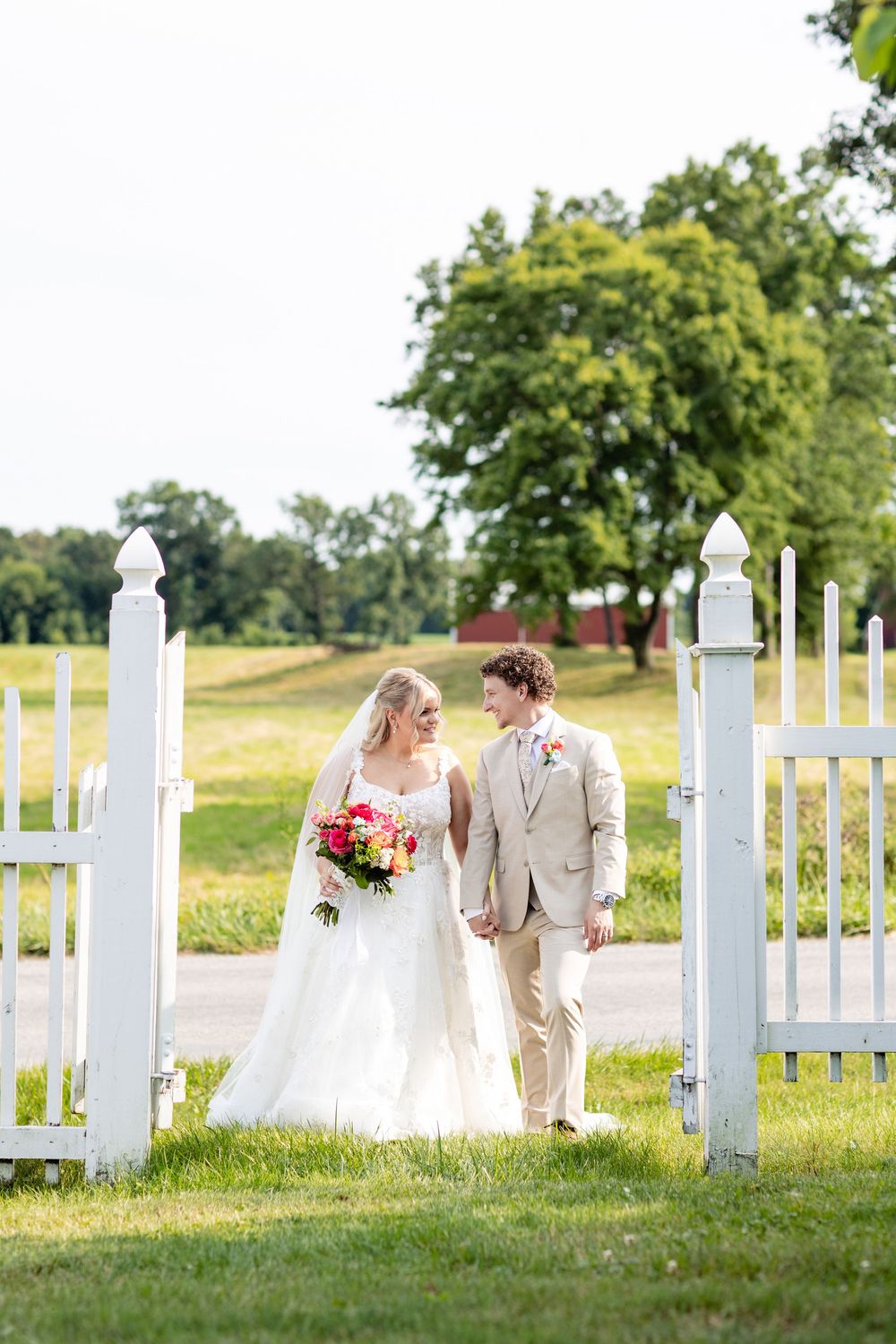 Bride and groom holding hands walking through white picket gate on green lawn with trees in background.