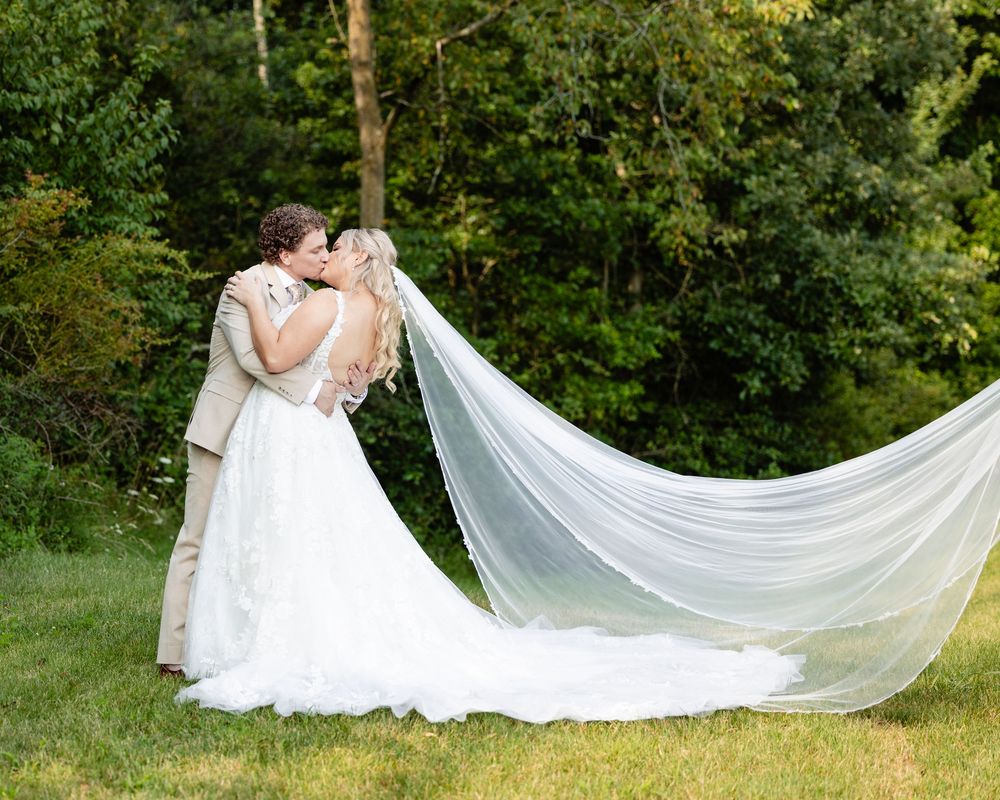 A bride and groom share a romantic kiss outdoors, with her long flowing veil billowing in the breeze against lush green trees.