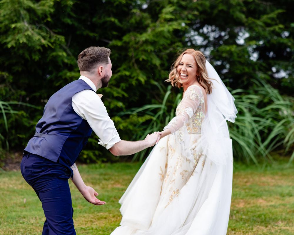 Joyful bride in lace gown and groom in navy suit dancing on green lawn during outdoor wedding celebration.